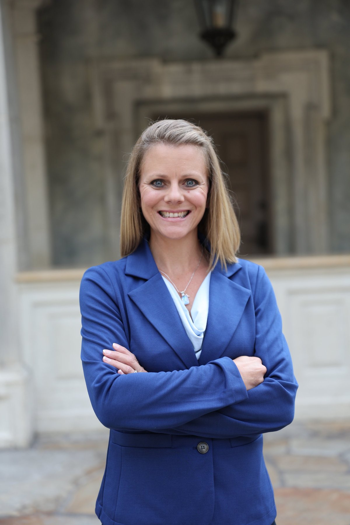 Professional woman with long blonde hair smiling at camera, wearing a blue blazer and white top, arms crossed, standing in front of a stone building entrance.