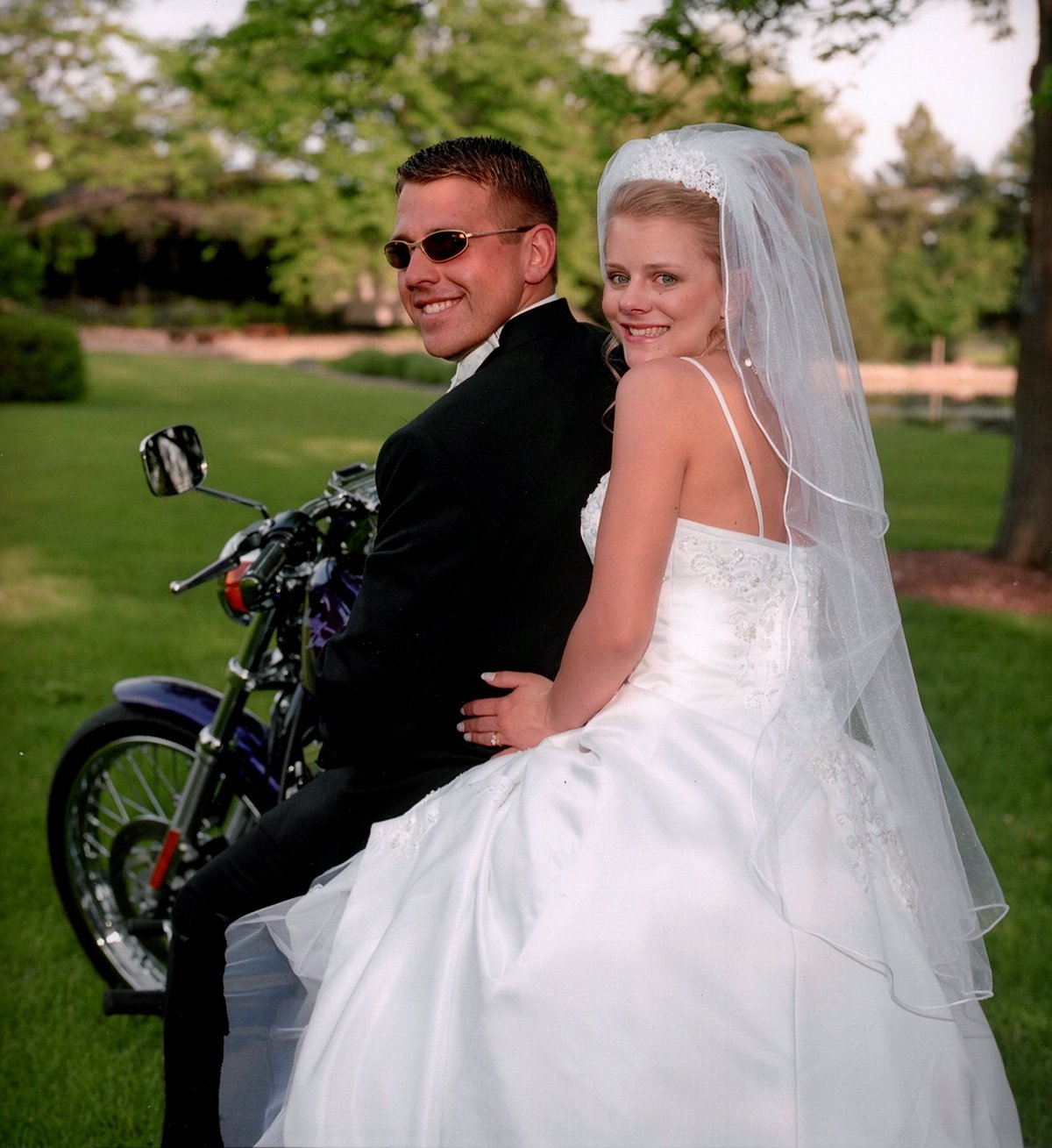 Bride and groom sitting on a motorcycle in a park, bride in white wedding dress and veil, groom in black suit and sunglasses